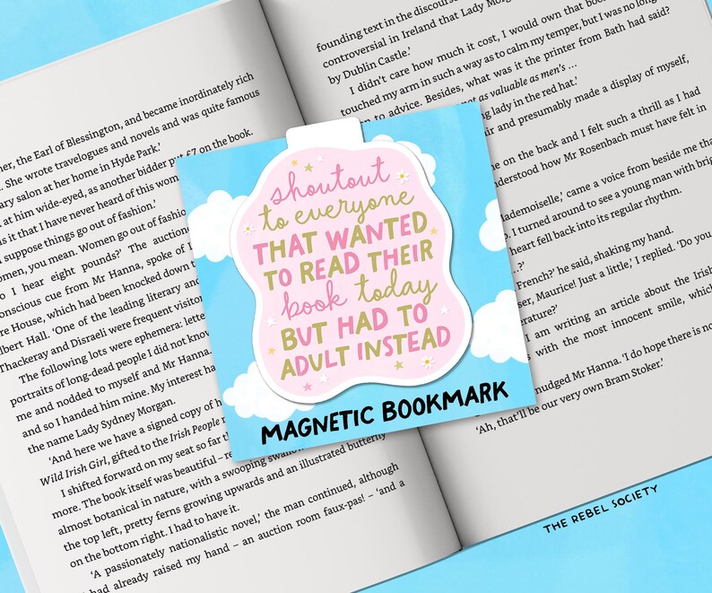 May include: A magnetic bookmark with a light blue background and a pink and white cloud-shaped design. The text reads "shoutout to everyone that wanted to read their book today but had to adult instead."