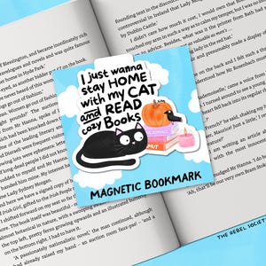 May include: A magnetic bookmark with a black cat illustration, a pumpkin, a crow, and a stack of books. The text reads "I just wanna stay HOME with my CAT and READ cozy Books". The bookmark is on an open book.