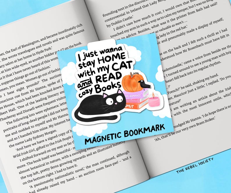 May include: A magnetic bookmark with a black cat illustration, a pumpkin, a crow, and a stack of books. The text reads "I just wanna stay HOME with my CAT and READ cozy Books". The bookmark is on an open book.
