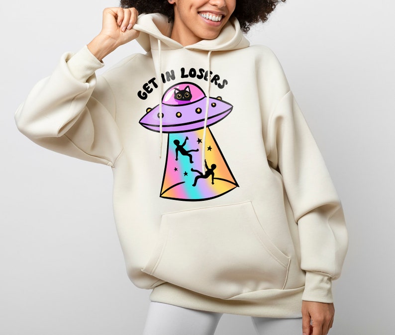 Puede incluir: Una sudadera con capucha de color crema con un gr&aacute;fico de un OVNI con un gato negro en su interior. El OVNI emite un rayo de luz de color arco&iacute;ris, y dos figuras de palo caen del cielo. El texto "GET IN LOSERS" est&aacute; impreso sobre el OVNI.
