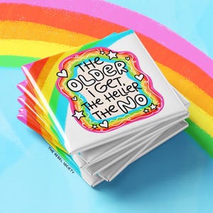May include: A stack of square white coasters featuring a colorful rainbow border and the black text: THE OLDER I GET, THE HELLER THE NO. The design includes small white stars and hearts, with the text THE REBEL SOCIETY printed in the bottom corner.