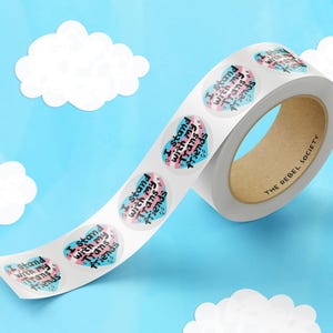 May include: Roll of stickers with a heart design and the text "I Stand with my Trans Friends" in black lettering on a blue and pink background. The roll is labeled "The Rebel Society".