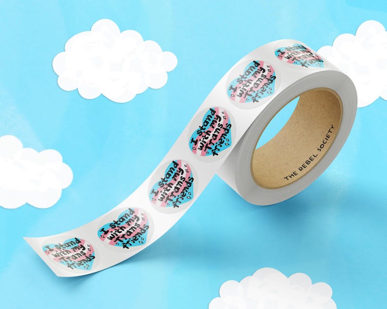 May include: Roll of stickers with a heart design and the text "I Stand with my Trans Friends" in black lettering on a blue and pink background. The roll is labeled "The Rebel Society".