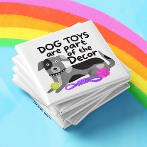 May include: A stack of white square coasters with a cartoon dog illustration and the text "DOG TOYS are part of the Decor". The dog is gray and white with a tennis ball and purple rope toy. The background features a rainbow.