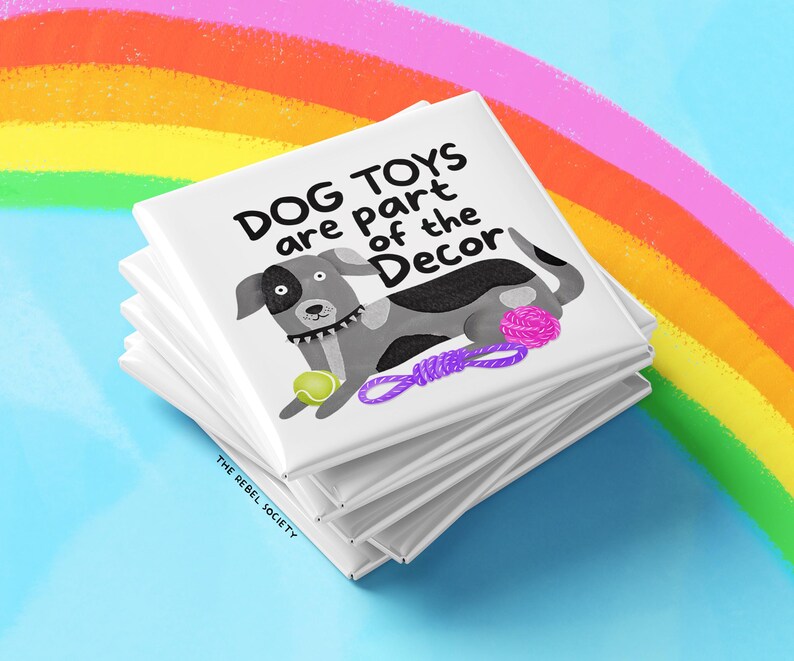 May include: A stack of white square coasters with a cartoon dog illustration and the text "DOG TOYS are part of the Decor". The dog is gray and white with a tennis ball and purple rope toy. The background features a rainbow.