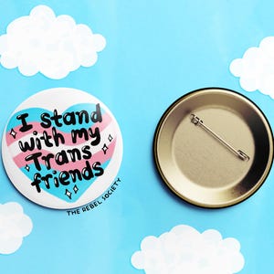 May include: A heart-shaped button with the text "I stand with my Trans friends" in black lettering on a pink, blue, and white transgender pride flag design. The button is on a blue background with white clouds. The back of the button is gold with a safety pin.
