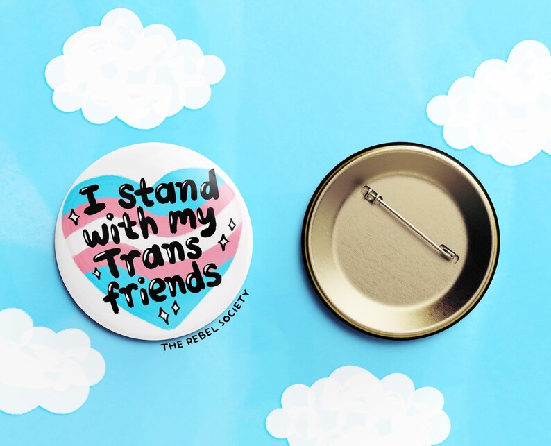 May include: A heart-shaped button with the text "I stand with my Trans friends" in black lettering on a pink, blue, and white transgender pride flag design. The button is on a blue background with white clouds. The back of the button is gold with a safety pin.