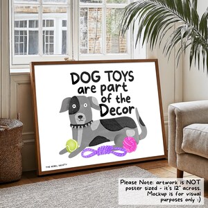 May include: A framed poster with the text "DOG TOYS are part of the Decor" and a cartoon dog illustration. The dog is gray and white, with a green tennis ball, a purple rope toy, and a pink yarn ball. The artwork is 12 inches across.