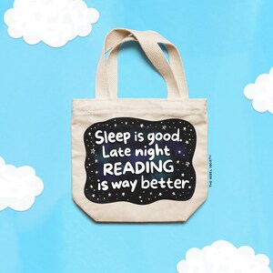 Pu&ograve; includere: Una borsa tote beige con la scritta "Sleep is good. Late night READING is way better." in un design in bianco e nero. La borsa ha due manici ed &egrave; ambientata su uno sfondo blu con ritagli di nuvole bianche.
