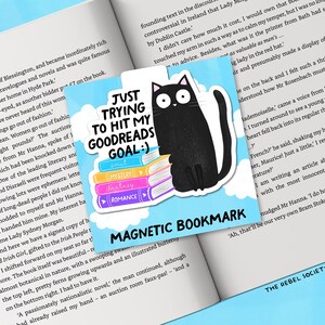 May include: A square magnetic bookmark with a black cat illustration sitting on a stack of colorful books. Text reads "JUST TRYING TO HIT MY GOODREADS GOAL :)" and "MAGNETIC BOOKMARK". The bookmark is on an open book.