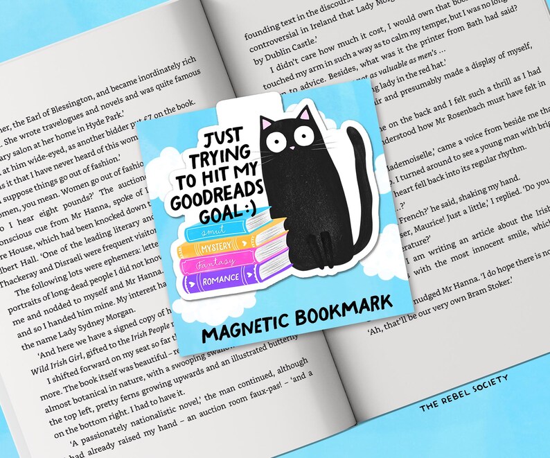 May include: A square magnetic bookmark with a black cat illustration sitting on a stack of colorful books. Text reads "JUST TRYING TO HIT MY GOODREADS GOAL :)" and "MAGNETIC BOOKMARK". The bookmark is on an open book.