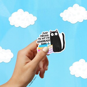 May include: A sticker featuring a black cat sitting next to a stack of books. The sticker has the text "JUST TRYING TO HIT MY GOODREADS GOAL :)" on a blue background with white clouds.