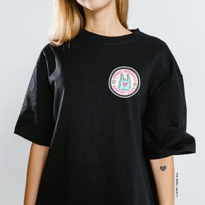 May include: Black short-sleeved t-shirt with a circular patch featuring a blue shopping bag with a pink heart, surrounded by text and floral designs. The patch reads "SMALL BUSINESS SUPPORT CLUB".