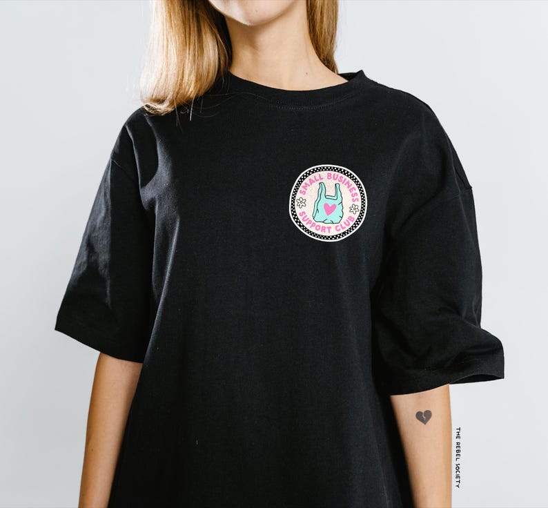 May include: Black short-sleeved t-shirt with a circular patch featuring a blue shopping bag with a pink heart, surrounded by text and floral designs. The patch reads "SMALL BUSINESS SUPPORT CLUB".