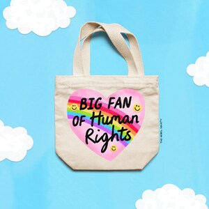 K&ouml;nnte beinhalten: Beige Canvas-Tragetasche mit einem herzf&ouml;rmigen Design mit dem Text "BIG FAN of Human Rights" in Schwarz und Regenbogenfarben. Die Tasche hat passende Griffe. Der Hintergrund ist hellblau mit wei&szlig;en Wolkenausschnitten.