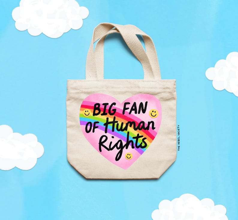 K&ouml;nnte beinhalten: Beige Canvas-Tragetasche mit einem herzf&ouml;rmigen Design mit dem Text "BIG FAN of Human Rights" in Schwarz und Regenbogenfarben. Die Tasche hat passende Griffe. Der Hintergrund ist hellblau mit wei&szlig;en Wolkenausschnitten.