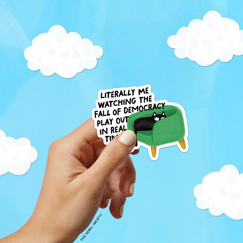 May include: A sticker featuring a black cat lounging on a green armchair, with the text "Literally me watching the fall of democracy play out in real time." The background is a blue sky with white clouds.