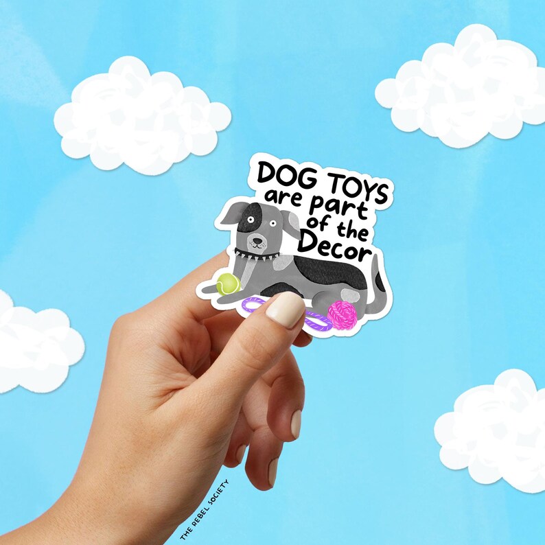 May include: A sticker featuring a cartoon dog with the text "DOG TOYS are part of the Decor." The dog is gray and white, with a tennis ball, a purple rope toy, and a pink ball. The background is a blue sky with white cloud cutouts.