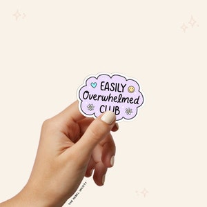 May include: A purple cloud-shaped sticker with the text "EASILY Overwhelmed CLUB" in black. A blue heart and a yellow smiley face are on either side of the text. There are small white flowers around the text.