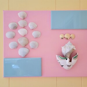 以下が含まれることがあります： ピンク色のキャンバスアート作品。白い貝殻がひし形に配置され、金色の貝殻のアクセントが3つ、水色の長方形のタイルが2つあります。大きな貝殻と割れた陶器の破片も含まれています。