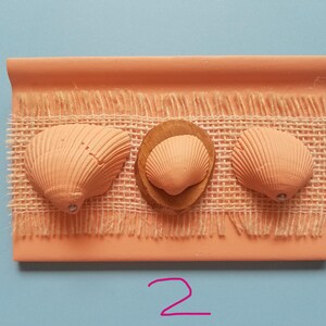 Puede incluir: Una pieza rectangular de color terracota con tres conchas marinas adjuntas. Las conchas marinas son de diferentes tamaños y están unidas a una tela tipo arpillera. El número "2" está escrito en rosa debajo.
