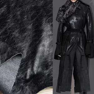 Puede incluir: Primer plano de un material de cuero negro texturizado con grano visible. A la derecha se muestra una gabardina de cuero negro con cinturón y elementos decorativos. El abrigo tiene un cuello de piel.