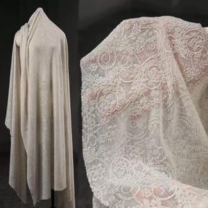 May include: Off-white fabric with a delicate floral lace pattern. The fabric is draped over a mannequin and also held up to show the intricate details of the lace. The fabric appears soft and lightweight, suitable for various craft projects.