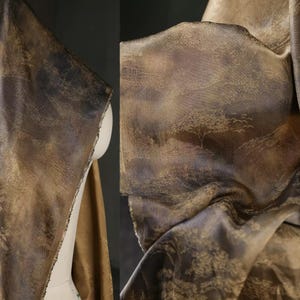 Può includere: Primo piano di un tessuto drappeggiato con un disegno a paesaggio. Il tessuto presenta un motivo di alberi e colline nelle tonalità del marrone, dell'oro e del grigio. Il materiale appare strutturato e ha una leggera lucentezza, suggerendo una qualità lussuosa.