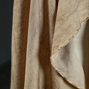 Puede incluir: Primer plano de una tela beige con una superficie texturizada y un borde sin rematar. La tela tiene un sutil patr&oacute;n de marcas marrones oscuras, creando una est&eacute;tica natural y terrosa. Los pliegues de la tela son visibles, a&ntilde;adiendo profundidad y dimensi&oacute;n a la imagen.