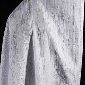 May include: A close-up of a white fabric with a textured, vertical ribbed pattern. The fabric is draped, with folds and shadows adding depth. The material appears soft and lightweight, suitable for clothing or home decor.