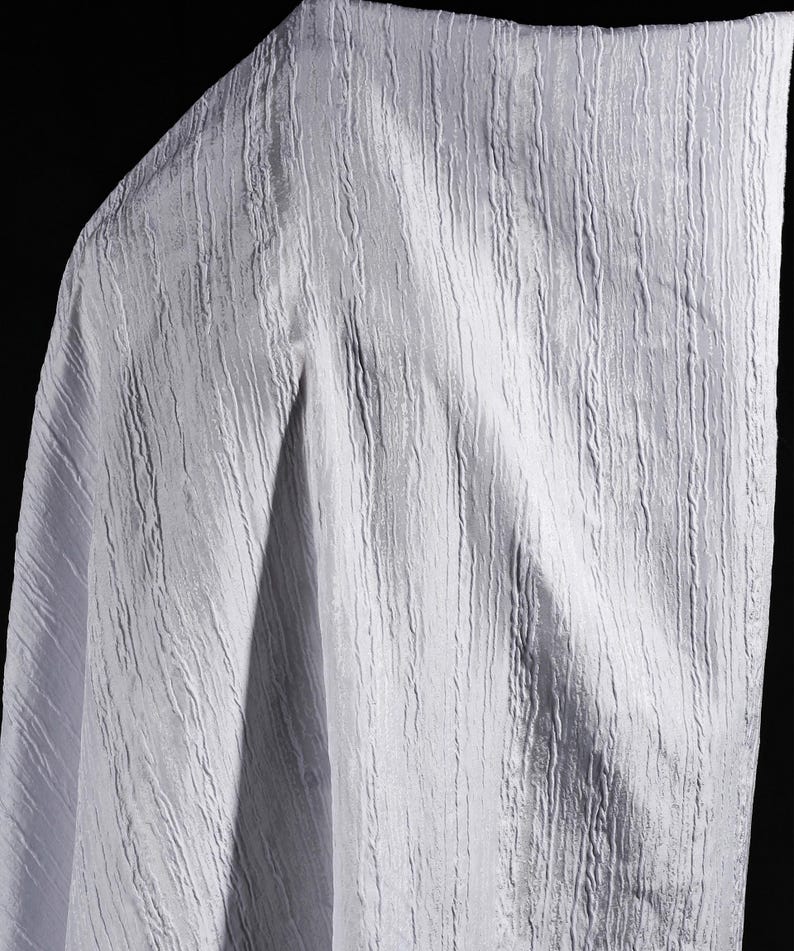 May include: A close-up of a white fabric with a textured, vertical ribbed pattern. The fabric is draped, with folds and shadows adding depth. The material appears soft and lightweight, suitable for clothing or home decor.
