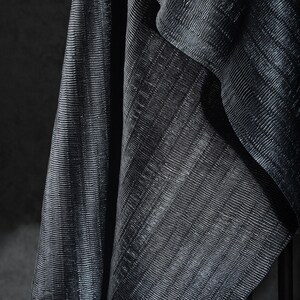 May include: Close-up of a dark, textured fabric with a subtle sheen. The material is draped, revealing diagonal lines and folds. The fabric's surface has a ribbed pattern, creating a play of light and shadow.