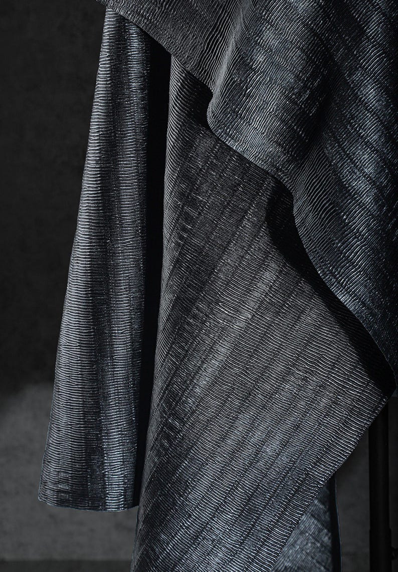 May include: Close-up of a dark, textured fabric with a subtle sheen. The material is draped, revealing diagonal lines and folds. The fabric's surface has a ribbed pattern, creating a play of light and shadow.
