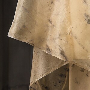 May include: Close-up of a layered, sheer fabric with a beige hue and dark gray mottled patterns. The fabric is folded, revealing multiple layers and a stitched edge. The material appears delicate and translucent.