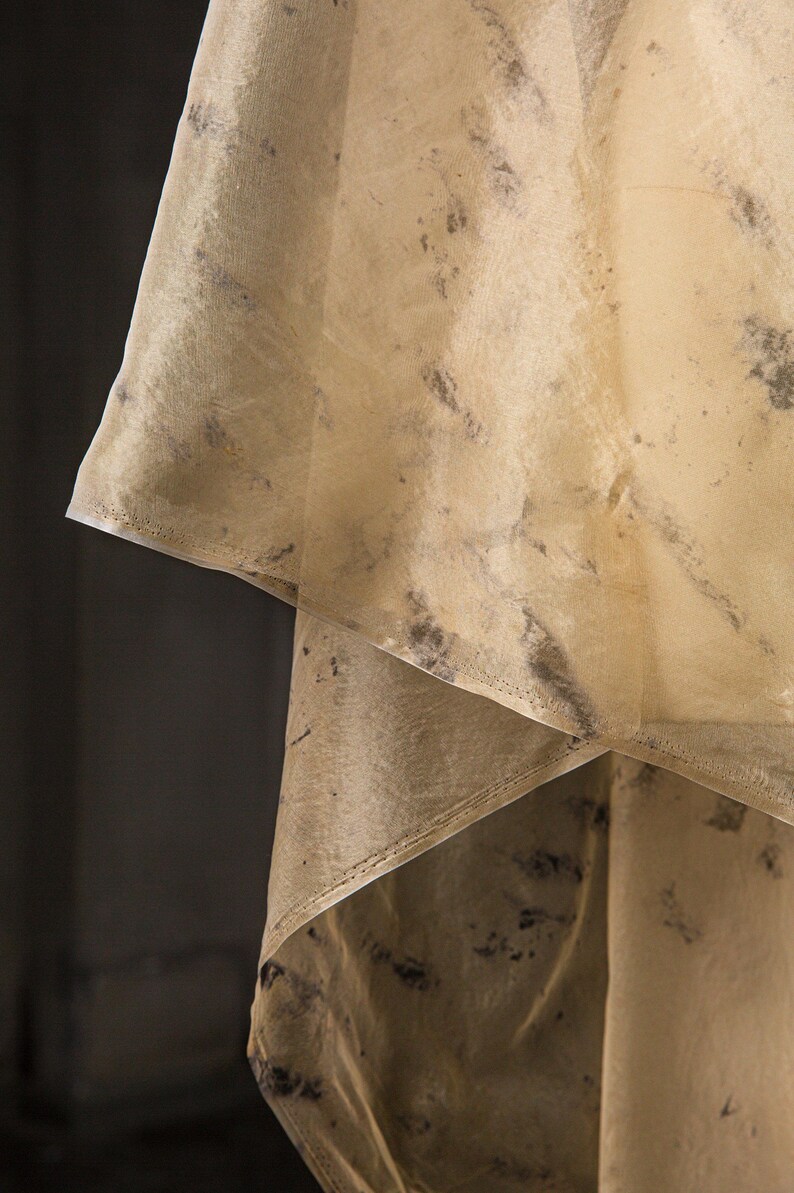 May include: Close-up of a layered, sheer fabric with a beige hue and dark gray mottled patterns. The fabric is folded, revealing multiple layers and a stitched edge. The material appears delicate and translucent.