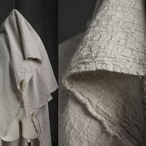 Può includere: Primo piano di un tessuto strutturato bianco sporco con un aspetto stropicciato. Il tessuto è drappeggiato, mostrando la sua consistenza e il modo in cui cade. Il colore neutro e la trama stropicciata sono le caratteristiche principali.