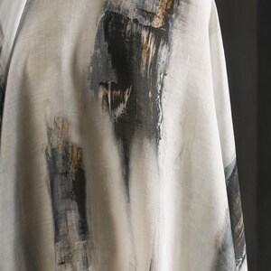 May include: Off-white fabric with an abstract design featuring vertical brushstrokes in black, grey, and gold. The fabric is draped, showcasing the texture and pattern. The overall aesthetic is artistic and modern.