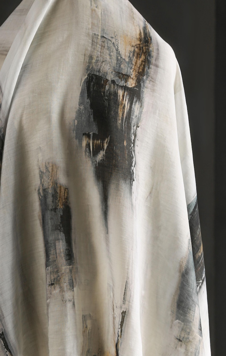 May include: Off-white fabric with an abstract design featuring vertical brushstrokes in black, grey, and gold. The fabric is draped, showcasing the texture and pattern. The overall aesthetic is artistic and modern.