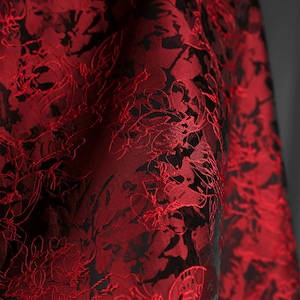 K&ouml;nnte beinhalten: Nahaufnahme eines luxuri&ouml;sen Stoffes mit floralem Muster. Der Stoff ist tiefrot mit schwarzen Akzenten, was einen auff&auml;lligen Kontrast erzeugt. Das florale Design ist detailliert und komplex und tr&auml;gt zur Eleganz des Stoffes bei.