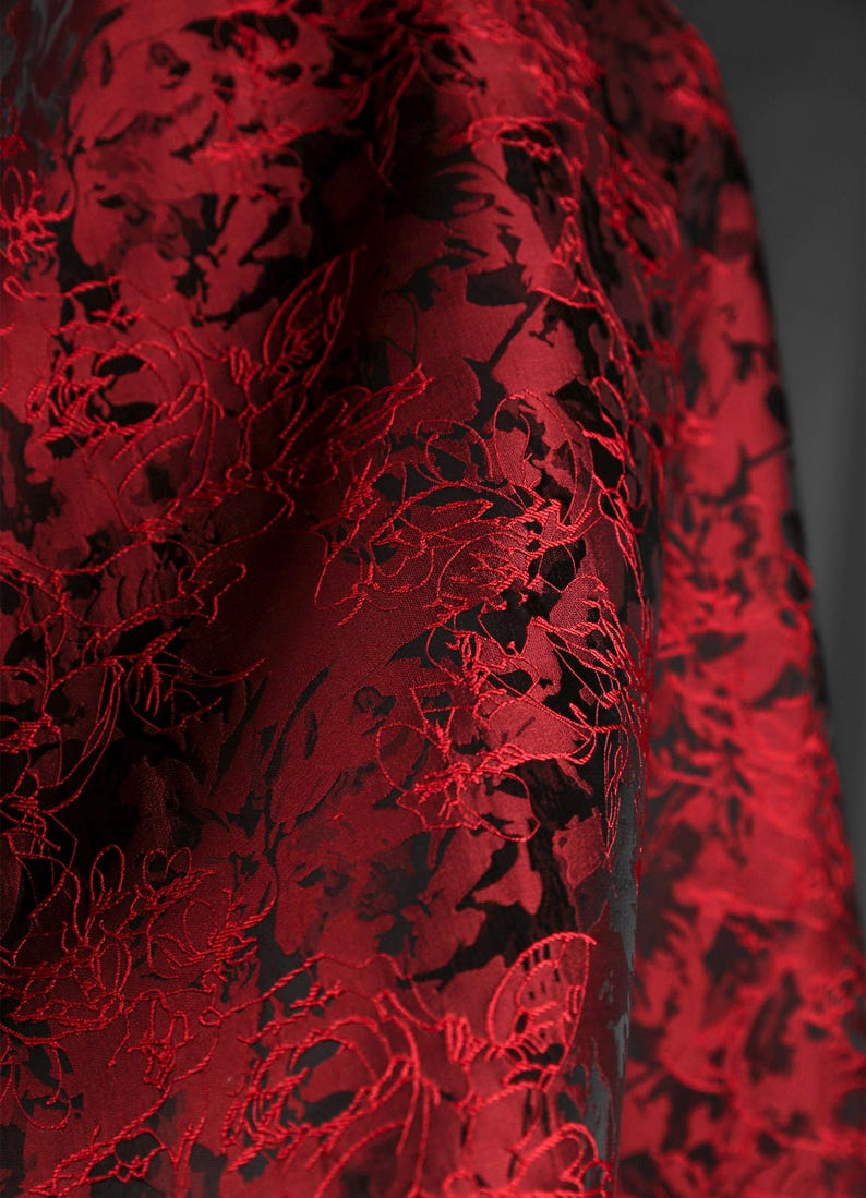 K&ouml;nnte beinhalten: Nahaufnahme eines luxuri&ouml;sen Stoffes mit floralem Muster. Der Stoff ist tiefrot mit schwarzen Akzenten, was einen auff&auml;lligen Kontrast erzeugt. Das florale Design ist detailliert und komplex und tr&auml;gt zur Eleganz des Stoffes bei.
