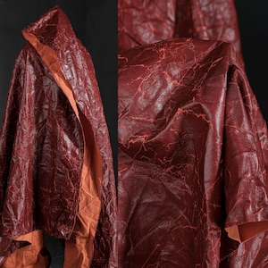 Könnte beinhalten: Ein burgunderfarbener Umhang mit einem strukturierten, rissigen Muster. Das Innenfutter hat eine kontrastierende orange Farbe. Der Umhang hat eine Kapuze und scheint aus einem flexiblen Material, möglicherweise Stoff oder Leder, zu bestehen.