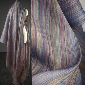 Op de afbeelding: Een gedrapeerde stof met verticale strepen in een spectrum van kleuren, waaronder blauw, paars, oranje en groen. De stof wordt getoond op een mannequin en in een close-up, waarbij de textuur en de drape worden getoond.