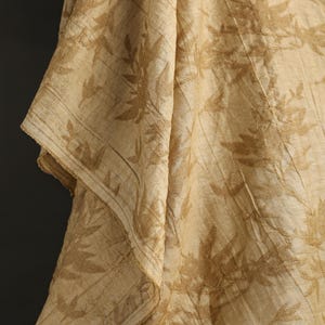 May include: Close-up of a beige fabric with a leaf pattern. The fabric is draped, showing folds and textures. The leaf design is in a slightly darker shade of beige, creating a subtle contrast. The edges of the fabric are visible.