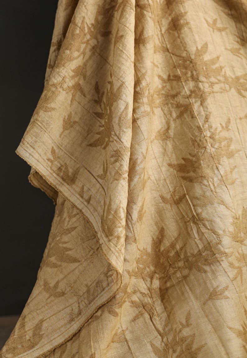 May include: Close-up of a beige fabric with a leaf pattern. The fabric is draped, showing folds and textures. The leaf design is in a slightly darker shade of beige, creating a subtle contrast. The edges of the fabric are visible.