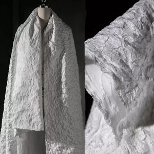 May include: A white textured fabric draped over a mannequin. The fabric has a raised, irregular pattern, creating a unique surface. The close-up reveals the intricate details of the material, highlighting its texture and design. The fabric appears to be a shawl or wrap.