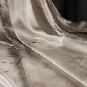 May include: Close-up of a flowing fabric with a marble-like pattern. The material is a blend of off-white, gray, and brown, creating a soft, textured appearance. The folds and curves of the fabric are highlighted by the light, giving it a luxurious feel.