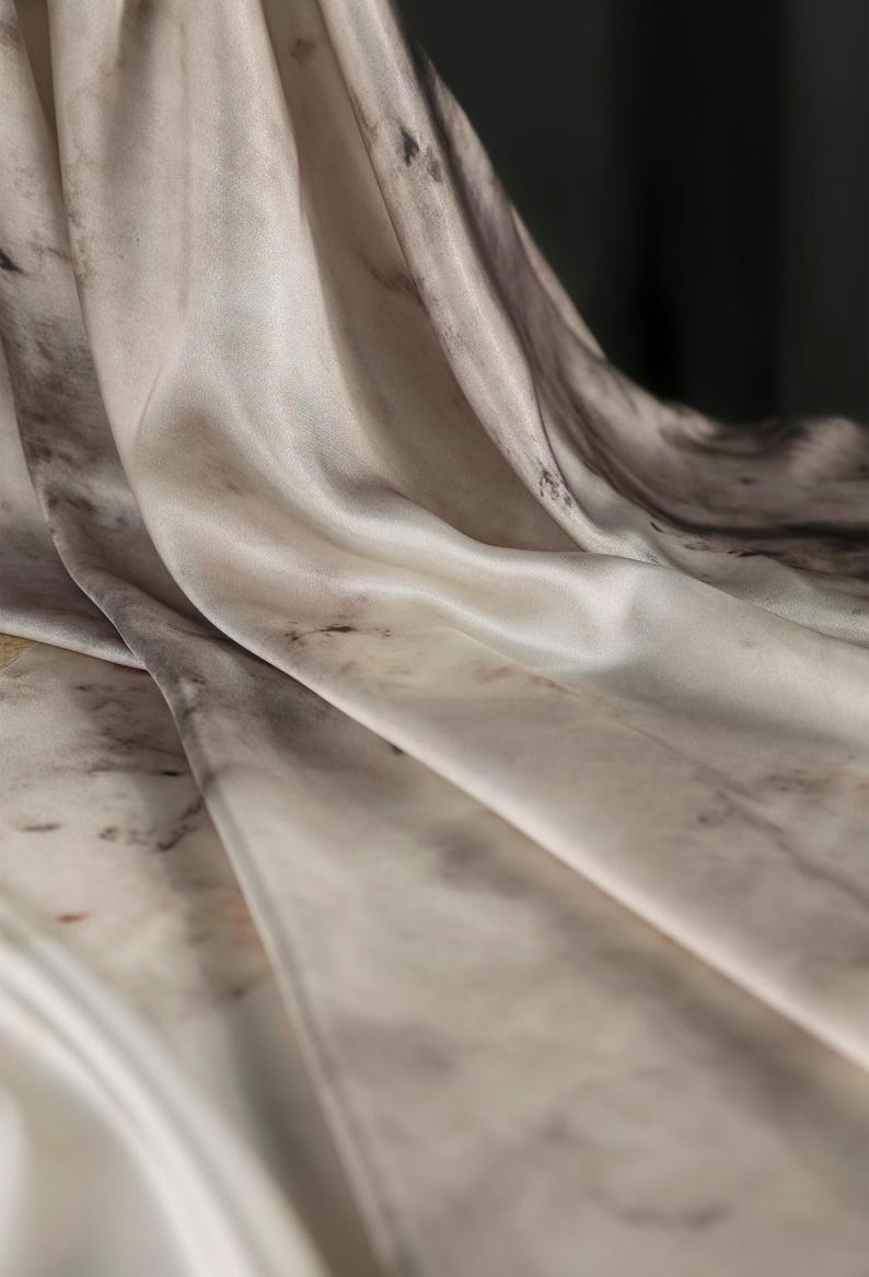 May include: Close-up of a flowing fabric with a marble-like pattern. The material is a blend of off-white, gray, and brown, creating a soft, textured appearance. The folds and curves of the fabric are highlighted by the light, giving it a luxurious feel.