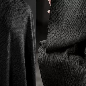 May include: Close-up of black fabric with a textured, scale-like pattern. The material appears to be draped, with folds and shadows adding depth. The fabric's surface has a subtle sheen, enhancing its visual texture.