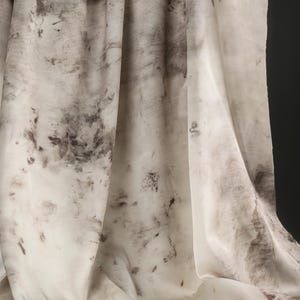 May include: Off-white fabric with a mottled pattern of dark gray and brown blotches. The fabric is draped, showing folds and shadows. The overall effect is organic and textured, resembling a watercolor painting.