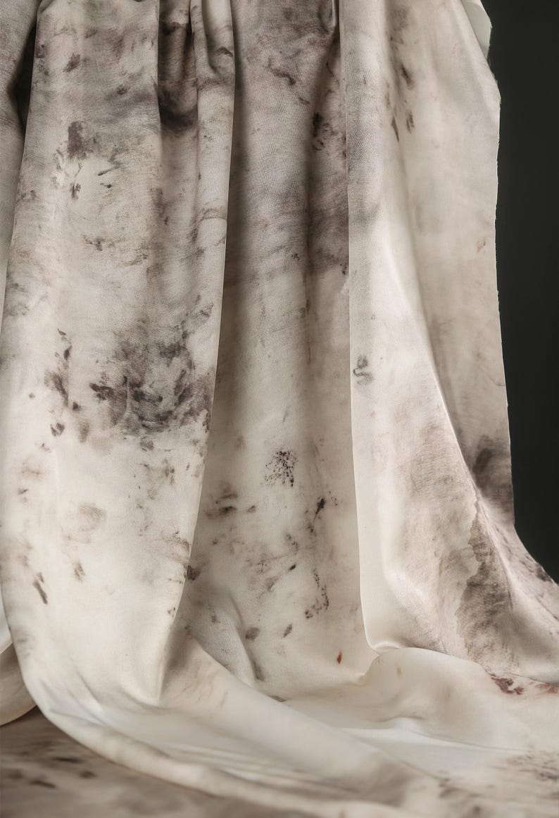 May include: Off-white fabric with a mottled pattern of dark gray and brown blotches. The fabric is draped, showing folds and shadows. The overall effect is organic and textured, resembling a watercolor painting.